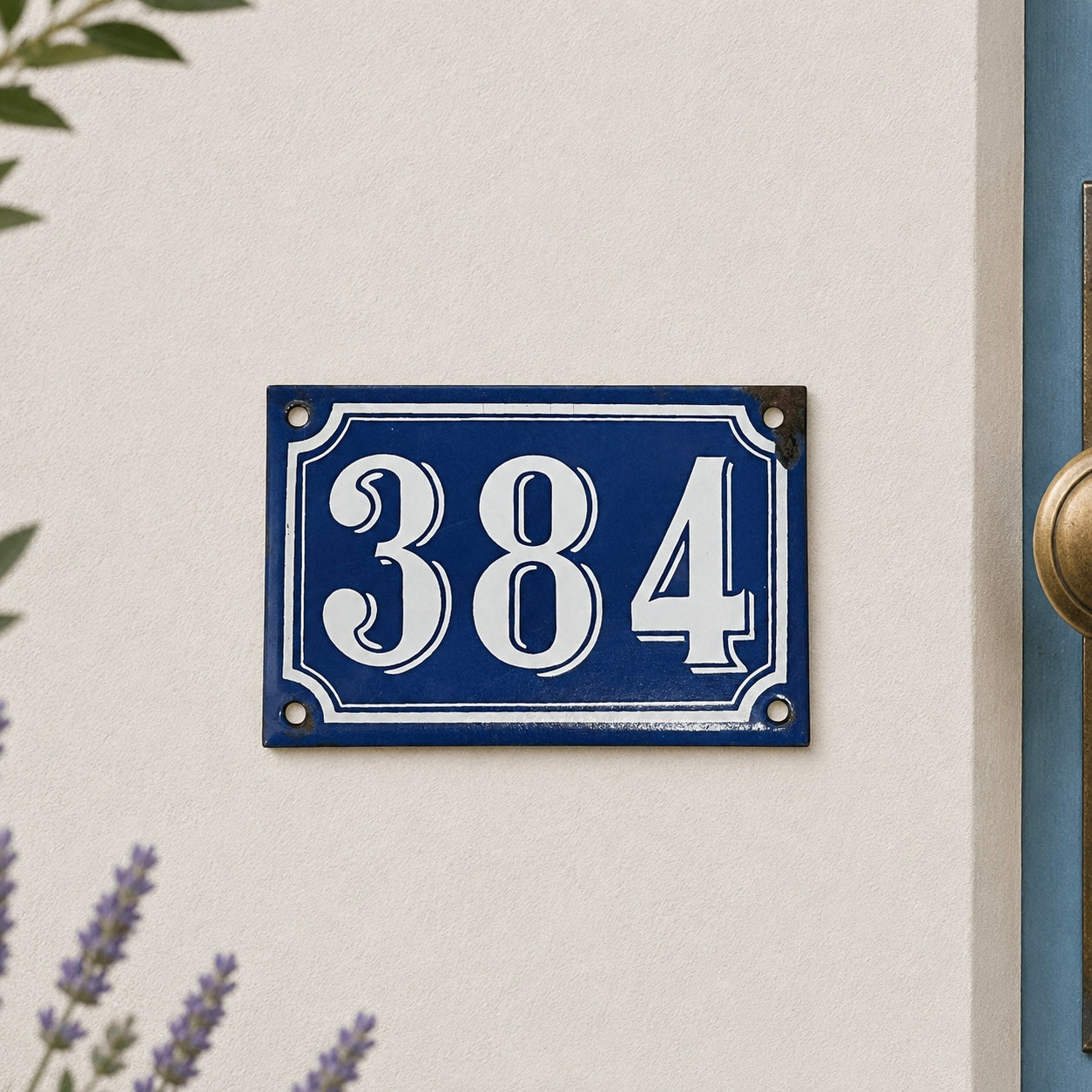 Vintage blue French enamel house number plaque displaying “384” in white numerals, mounted on a cream textured exterior wall beside a pale blue door with brass hardware. Soft natural light, subtle garden foliage and lavender flowers frame the scene, creating a charming French country house entrance