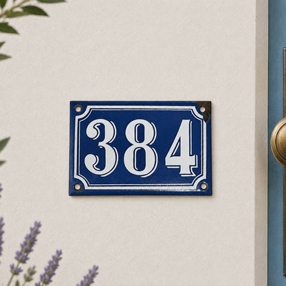 Vintage blue French enamel house number plaque displaying “384” in white numerals, mounted on a cream textured exterior wall beside a pale blue door with brass hardware. Soft natural light, subtle garden foliage and lavender flowers frame the scene, creating a charming French country house entrance
