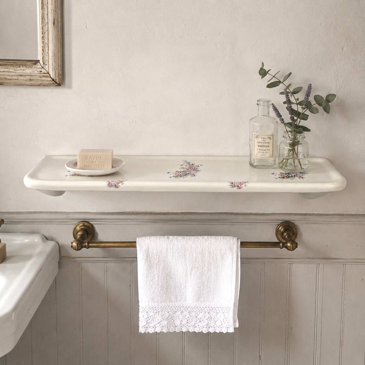 A white ceramic wall-mounted shelf with a delicate floral pattern, styled with a small dish, soap bar, and a glass bottle with eucalyptus. Below the shelf, a brass towel bar holds a white lace-trimmed towel in a bathroom setting with a white paneled wall.