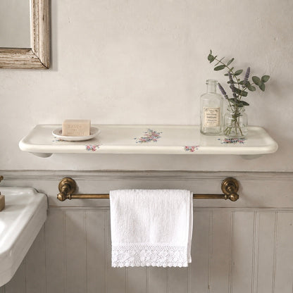 A white ceramic wall-mounted shelf with a delicate floral pattern, styled with a small dish, soap bar, and a glass bottle with eucalyptus. Below the shelf, a brass towel bar holds a white lace-trimmed towel in a bathroom setting with a white paneled wall.