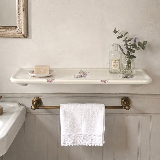 A white ceramic wall-mounted shelf with a delicate floral pattern, styled with a small dish, soap bar, and a glass bottle with eucalyptus. Below the shelf, a brass towel bar holds a white lace-trimmed towel in a bathroom setting with a white paneled wall.