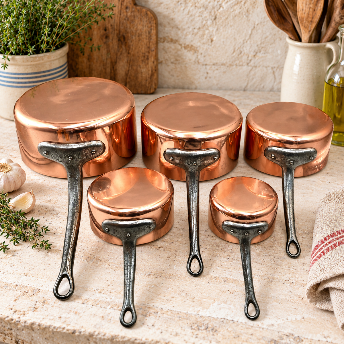 A set of copper cookware pots and pans with dark metal handles arranged on a stone countertop in a kitchen setting, with garlic, herbs, a cutting board, and a striped towel visible.