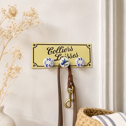 A decorative wall-mounted coat rack with the words 'Colliers Laisses' in black script on a pale yellow panel. The rack has three round ceramic hooks with blue and white floral patterns and holds brown leather straps and a brass clasp.