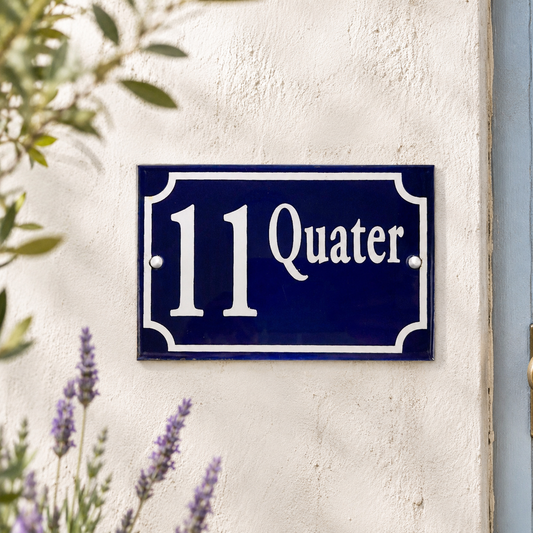 Blue enamel French-style house number plaque displaying the number 11 in white, mounted on a cream rendered exterior wall with visible fixing screws. Soft-focus lavender flowers and leafy branches frame the sign in natural daylight.