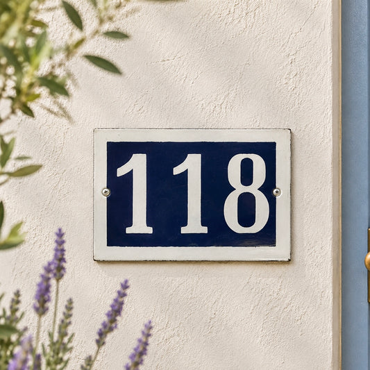 House number '118' on a blue plaque attached to a white wall with plants in the foreground.