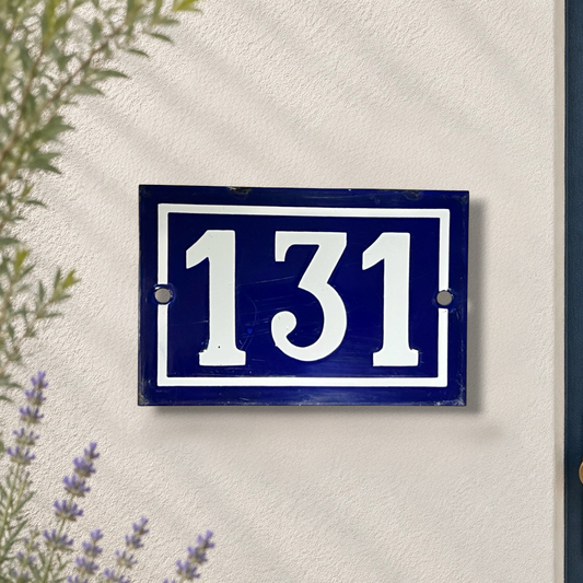 French vintage enamel house number 131, white number on a blue background, mounted on a pale stone wall with a lavender plant and the edge of a door at the side
