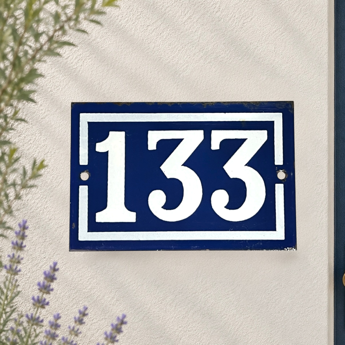 A rectangular navy blue house number plaque with white lettering reading '133', mounted on a textured off-white wall with soft leaf shadows and small plants visible at the edges.