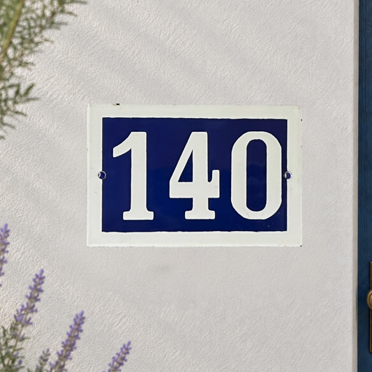 A rectangular house number plaque reading 140 in white on a dark blue background, mounted on a textured off-white wall with small plants visible at the edges.