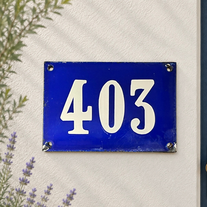 A rectangular blue French house number plaque with white numerals '403' mounted on a textured off-white wall, with green leaves and purple flowers partially visible in the foreground.