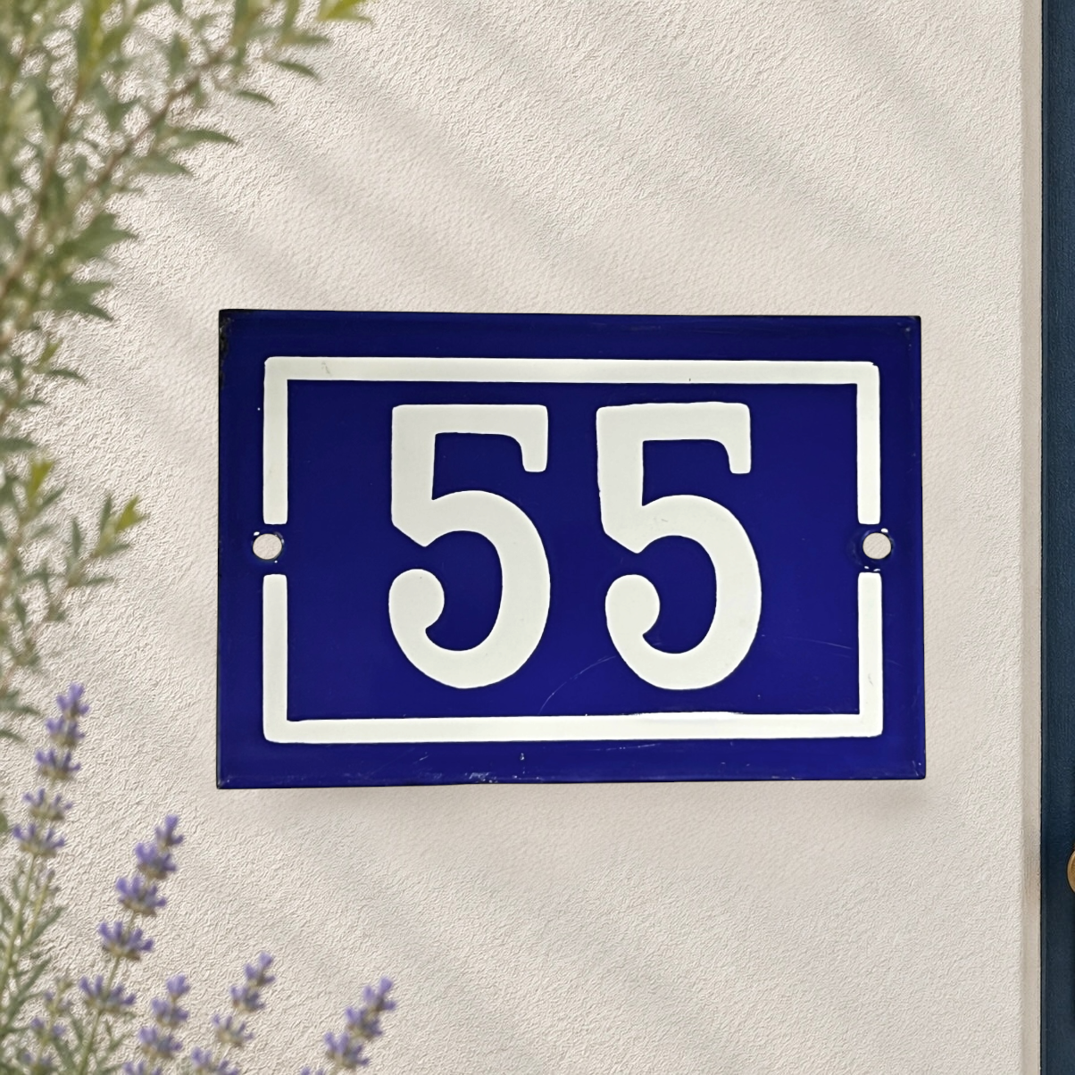 A rectangular blue house number plaque with white border and the numbers 55, mounted on a textured white stucco wall with some greenery and lavender visible at the edges.