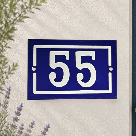 A rectangular blue house number plaque with white border and the numbers 55, mounted on a textured white stucco wall with some greenery and lavender visible at the edges.