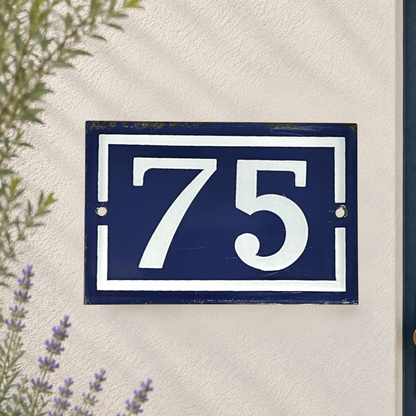A rectangular navy blue house number plaque with white lettering and border details number 75, mounted on a textured off-white wall with soft leaf shadows and small purple flowers in the foreground.