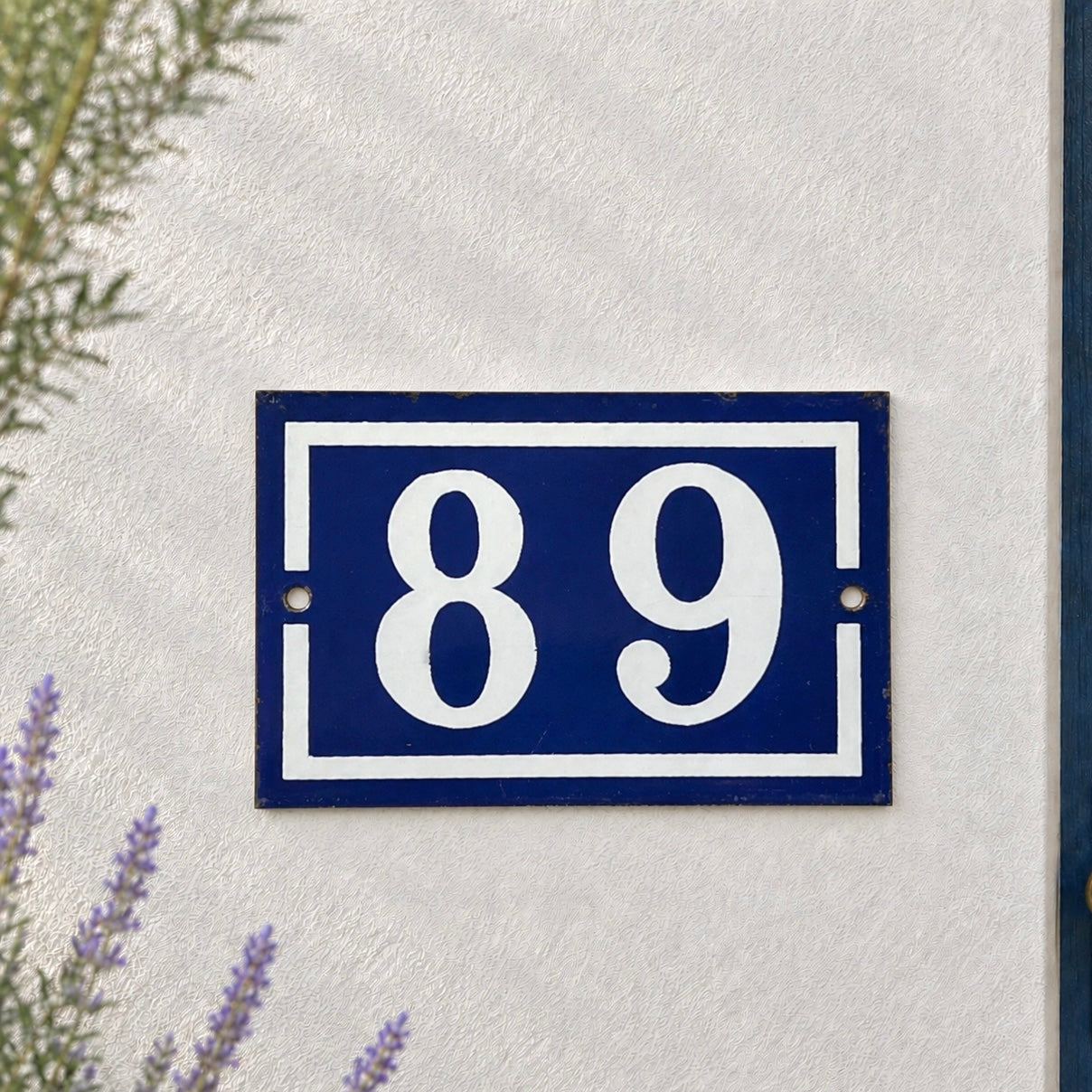 A rectangular blue house number plaque with white numerals '89' and a white border, mounted on a textured white wall next to a blue door frame.