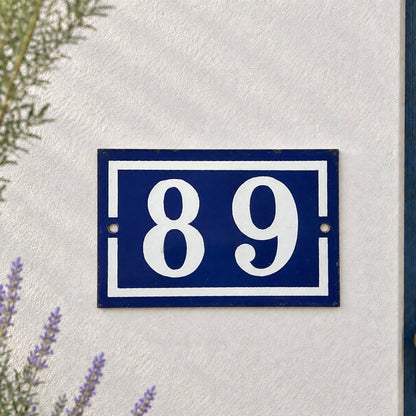 A rectangular blue house number plaque with white numerals '89' and a white border, mounted on a textured white wall next to a blue door frame.