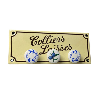 A vintage-style cream enamel pin or plaque featuring the French text 'Colliers Laisses' in dark brown script, with three small decorative ceramic buttons below: two white with blue floral wreath designs and one white with a blue flower and green stem. The item is photographed on a clean white background.