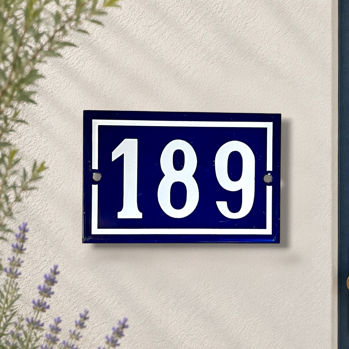 A rectangular navy blue house number plaque with white lettering reading '189', mounted on a textured off-white wall. Green leaves and small purple flowers are visible at the edges of the frame.