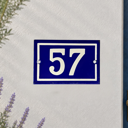 A blue and white French-style house number plaque reading '57' mounted on a textured white stucco wall, with small green and purple plant sprigs visible at the edges.