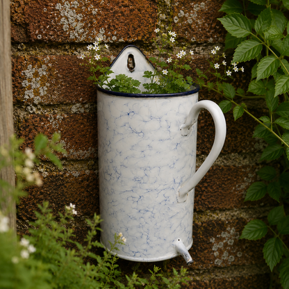 A white enamel  planter with blue marbled veins, a dark rim, and a white handle, shown holding green plants against a rustic brick wall.