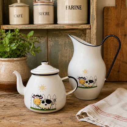 A white enamel teapot and matching pitcher set with black metal handles and spouts, decorated with a colourful cartoon cow and sun motif, displayed on a rustic wooden table with a potted plant and canisters in the background.