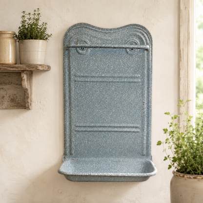 A speckled slate blue wall-mounted utensil rack with a curved top and integrated tray, shown mounted on a textured cream wall with potted plants nearby.