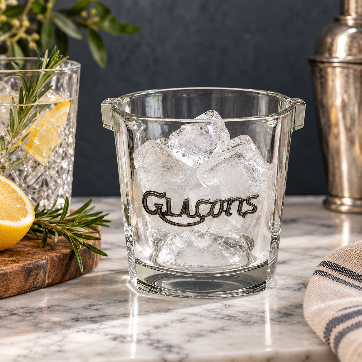A clear glass ice cube bucket with side handles and the word 'Glacons' printed in dark script, filled with ice on a marble countertop. A cocktail shaker, another glass with lemon and rosemary, and a striped cloth are visible in the background.