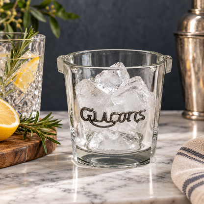 A clear glass ice cube bucket with side handles and the word 'Glacons' printed in dark script, filled with ice on a marble countertop. A cocktail shaker, another glass with lemon and rosemary, and a striped cloth are visible in the background.