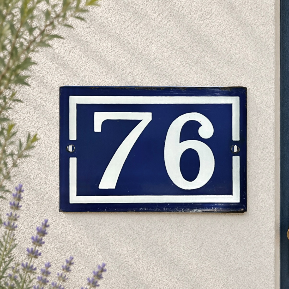 A rectangular navy blue house number plaque with white lettering reading '76', mounted on a textured off-white wall with some greenery and lavender visible at the edges.