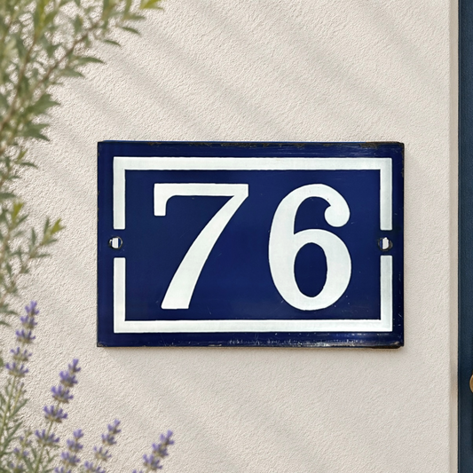A rectangular navy blue house number plaque with white lettering reading '76', mounted on a textured off-white wall with some greenery and lavender visible at the edges.