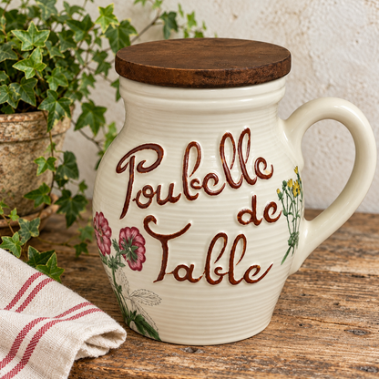 A cream-colored ceramic table jar with a dark wooden lid, featuring the French text 'Poubelle de Table' in brown script and pink floral illustrations. The jar is styled on a rustic wooden table next to a striped cloth napkin and green ivy.