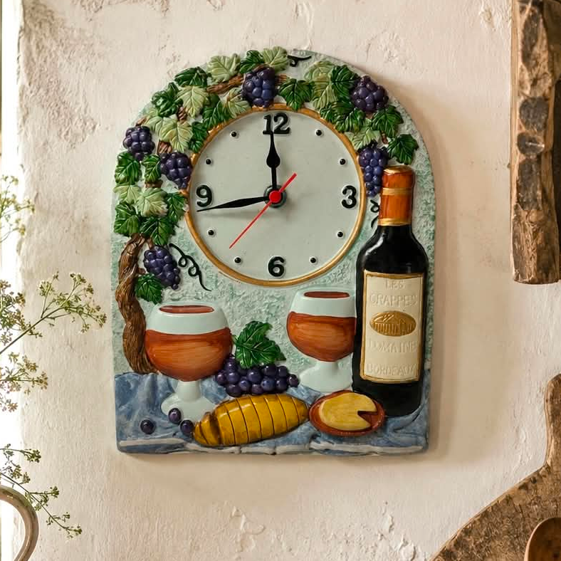 A decorative wall clock shaped like a rustic wine-themed plaque, featuring a white clock face surrounded by green leaves, purple grapes, a wine bottle, two wine glasses, and a sliced loaf of bread, mounted on a textured off-white wall.