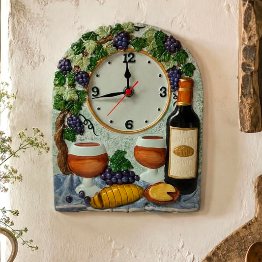 A decorative wall clock shaped like a rustic wine-themed plaque, featuring a white clock face surrounded by green leaves, purple grapes, a wine bottle, two wine glasses, and a sliced loaf of bread, mounted on a textured off-white wall.