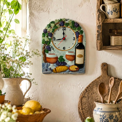 A decorative wall clock with a rustic French village theme, featuring grapes, wine bottles, bread, and cups on a blue background, mounted on a textured white wall in a cosy kitchen setting.