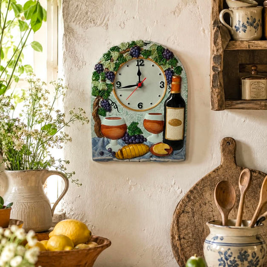 A decorative wall clock with a rustic French village theme, featuring grapes, wine bottles, bread, and cups on a blue background, mounted on a textured white wall in a cosy kitchen setting.