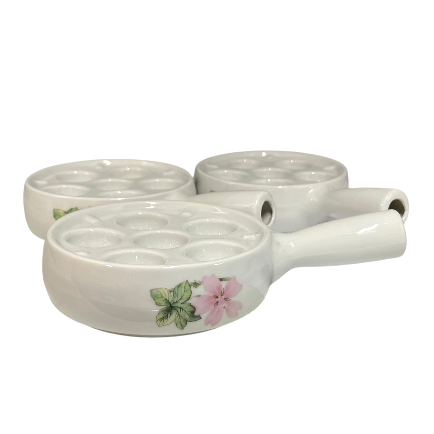 Three white ceramic egg plates with floral designs, featuring a pink flower and green leaves on the front poacher, set against a plain white background.
