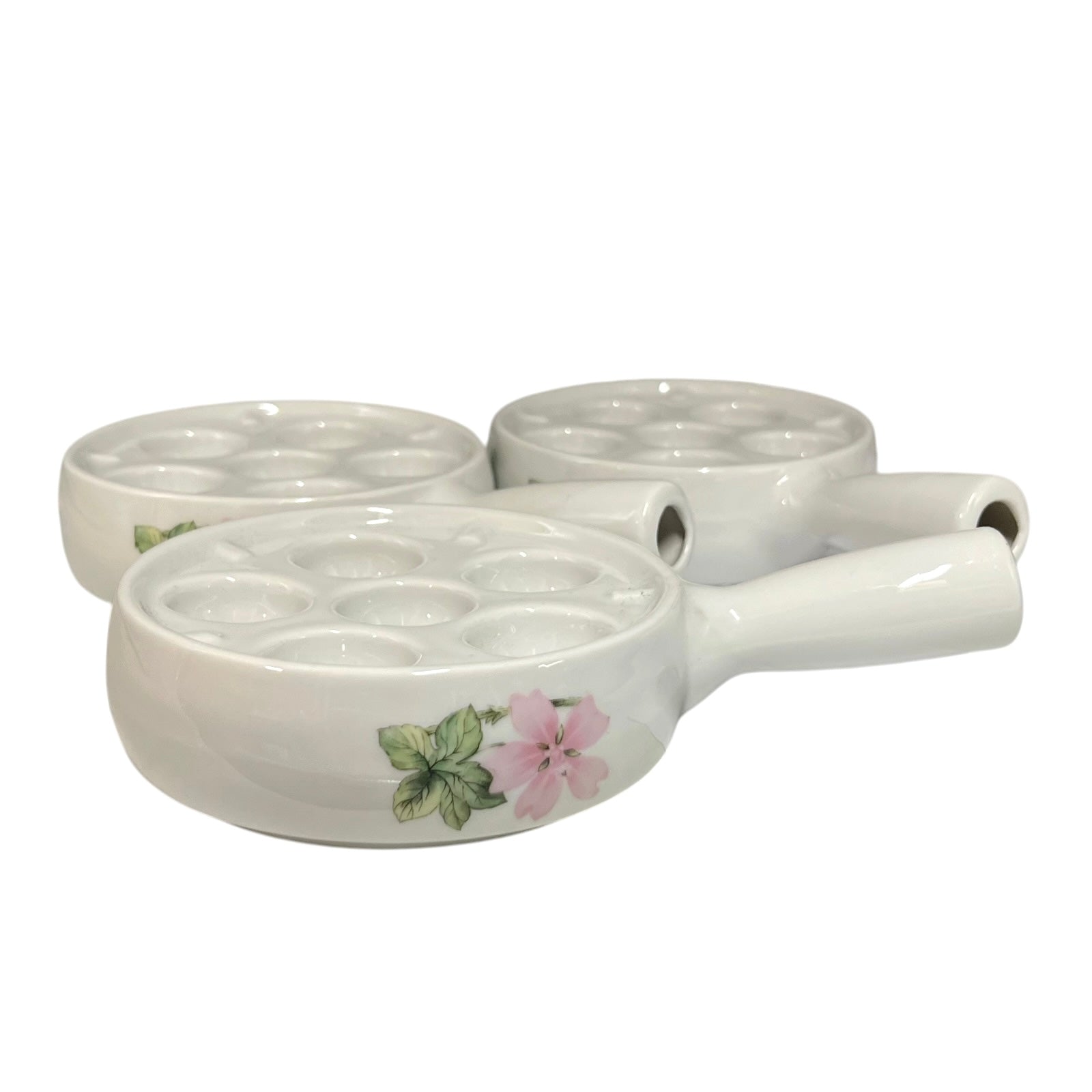 Three white ceramic egg plates with floral designs, featuring a pink flower and green leaves on the front poacher, set against a plain white background.