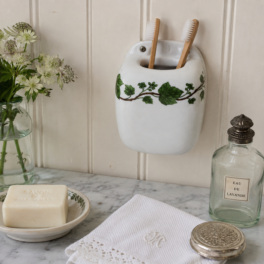 A white ceramic toothbrush holder with a green ivy vine design, mounted on a cream panelled wall above a marble countertop. The holder contains two wooden-handled toothbrushes.