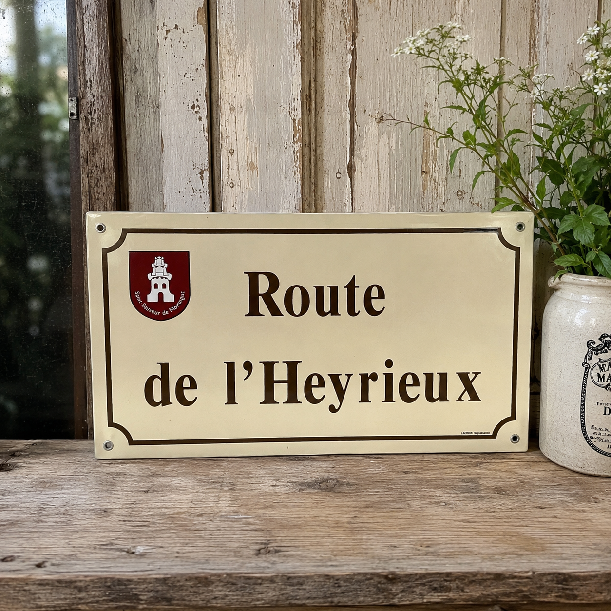 A rectangular cream-colored metal street sign with black lettering reading 'Route de l’Heyrieux' and a small red-and-white crest in the upper left, displayed on a rustic wooden surface against a weathered wood plank wall, with a white ceramic jar and greenery beside it.