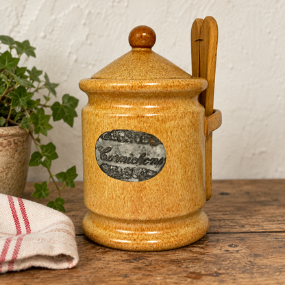 A glazed ceramic cornichon jar with a wooden handle and wooden spoon, displayed on a rustic wooden table with a potted plant and striped cloth in the background.