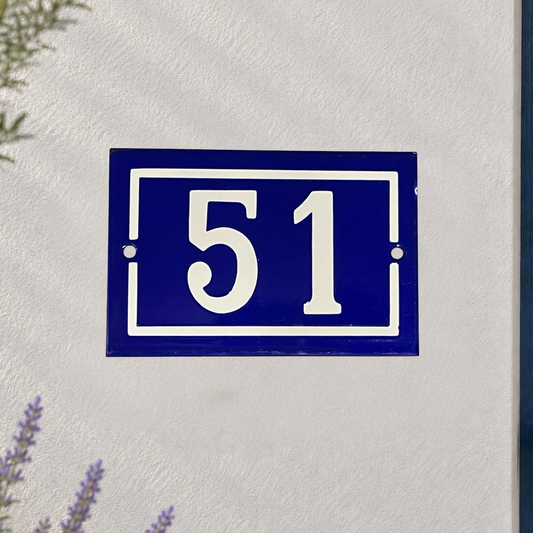 A blue French-style house number plaque with white numerals '51' mounted on a textured white wall, with small green and purple plants partially visible at the edges.