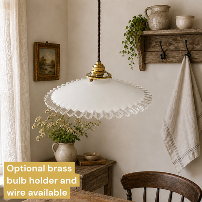 A white glass pendant light with a scalloped ruffled edge hangs over a rustic dining table in a cosy room. A brass bulb holder and cord are visible, with a text overlay stating 'Optional brass bulb holder and wire available'.