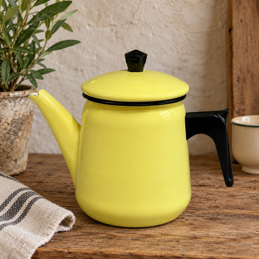 A bright yellow enamel coffee pot with black accents sits on a rustic wooden table, styled with a potted olive plant and a striped cloth in a warm, cosy setting.