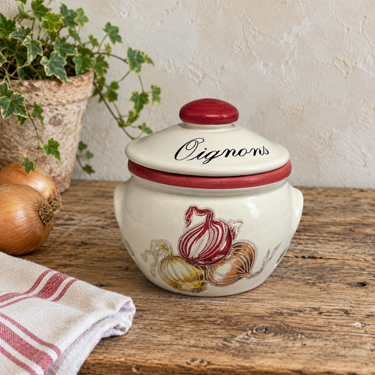 Vintage French ceramic onion storage jar with lid, displayed on a rustic wooden farmhouse kitchen table against a softly textured cream wall. The traditional cream glazed Oignons pot features burgundy-red detailing, a matching red lid knob, elegant black French lettering and raised decorative illustrations of red, yellow and brown onions. A rustic potted ivy plant, fresh onions and a folded white tea towel with red stripes complete the French country kitchen setting. 