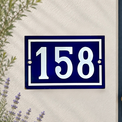 A rectangular dark blue house number plaque with white numerals '158' and white border details, mounted on a textured off-white wall with some greenery visible at the edges.
