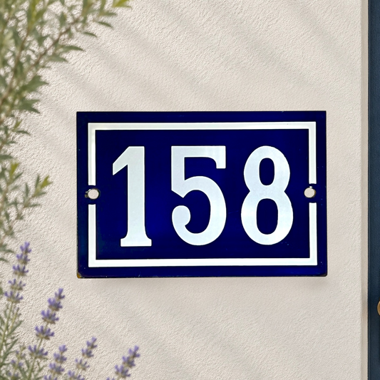 A rectangular dark blue house number plaque with white numerals '158' and white border details, mounted on a textured off-white wall with some greenery visible at the edges.