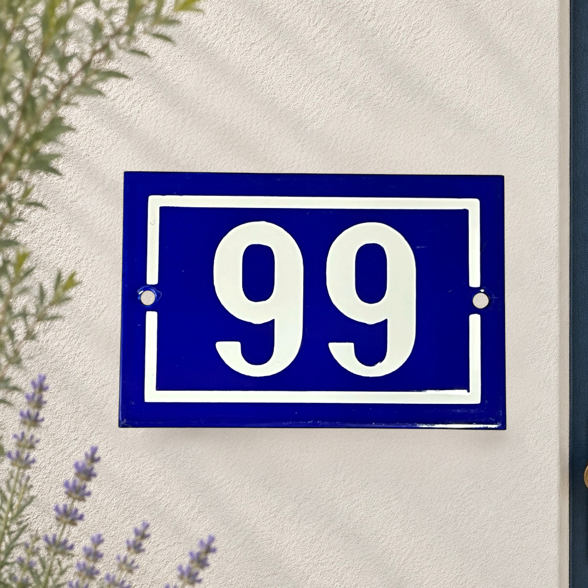 A classic blue and white French house number plaque showing '99' mounted on a textured white stucco wall, with soft shadows and a hint of greenery in the corners.