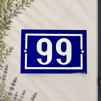 A classic blue and white French house number plaque showing '99' mounted on a textured white stucco wall, with soft shadows and a hint of greenery in the corners.