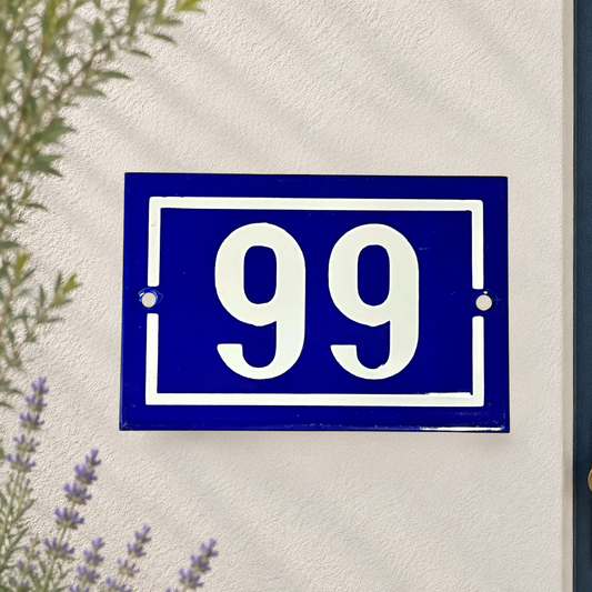 A classic blue and white French house number plaque showing '99' mounted on a textured white stucco wall, with soft shadows and a hint of greenery in the corners.