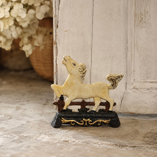 A decorative horse figurine in a cream and gold finish, standing on a dark base with ornate gold detailing, set against a rustic white wooden door and stone floor.