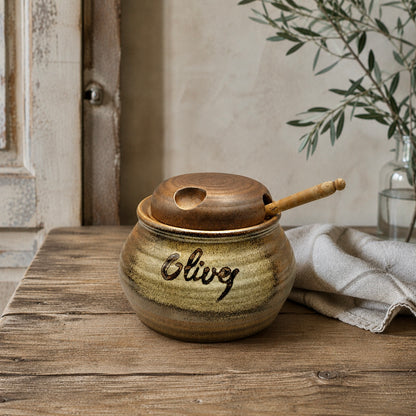 A ceramic jar with a lid and wooden handle sits on a wooden surface, adorned with the word 'Olive', perfect for adding charm to a farmhouse or country cottage.