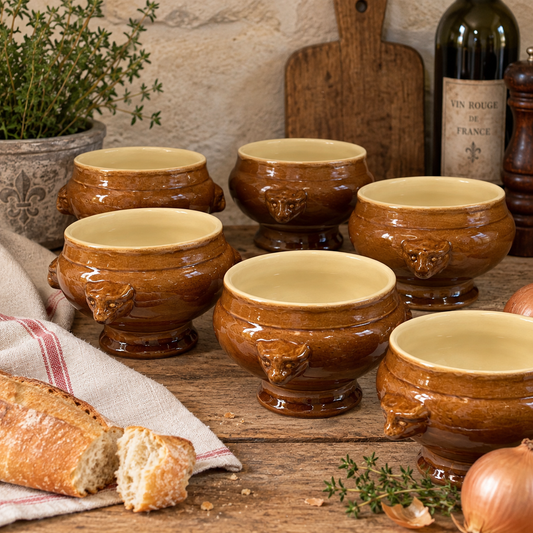 Five brown glazed ceramic soup bowls with animal-head handles are arranged on a rustic wooden table with bread, onions, herbs, a cutting board, and a wine bottle in the background.